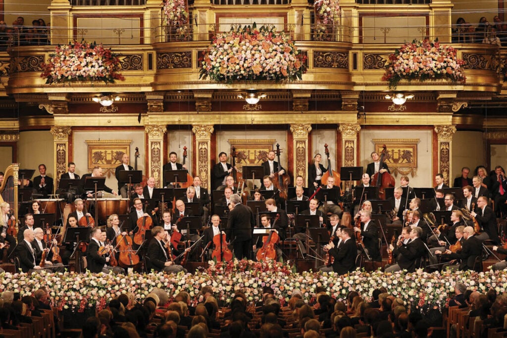 A fotografia mostra a Orquestra Filarmónica de Viena a atuar num palco elegante, com músicos em trajes formais a tocar vários instrumentos. A orquestra está posicionada à frente de um fundo dourado e decorado com flores, enquanto o maestro está de costas, conduzindo a performance. O público é visível nas primeiras filas.