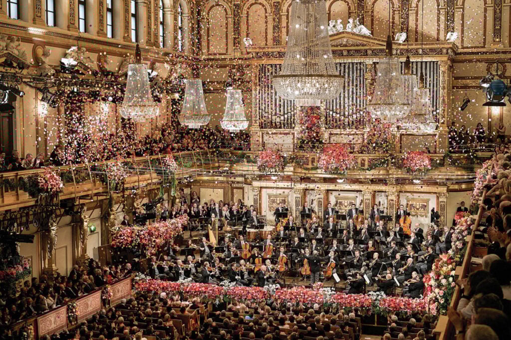 Orquestra filarmónica a actuar numa sala de concertos clássica ricamente decorada, com público nas galerias e confettis a cair do tecto. A Orquestra Filarmónica de Viena Rolex