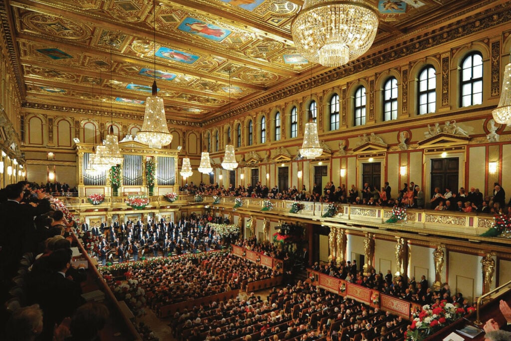 Vista geral de uma sala de concertos clássica ornamentada, com orquestra em palco, público nas galerias e grandes lustres de cristal. Orquestra Filarmónica de Viena no Salão Dourado do histórico Musikverein | Foto: Rolex