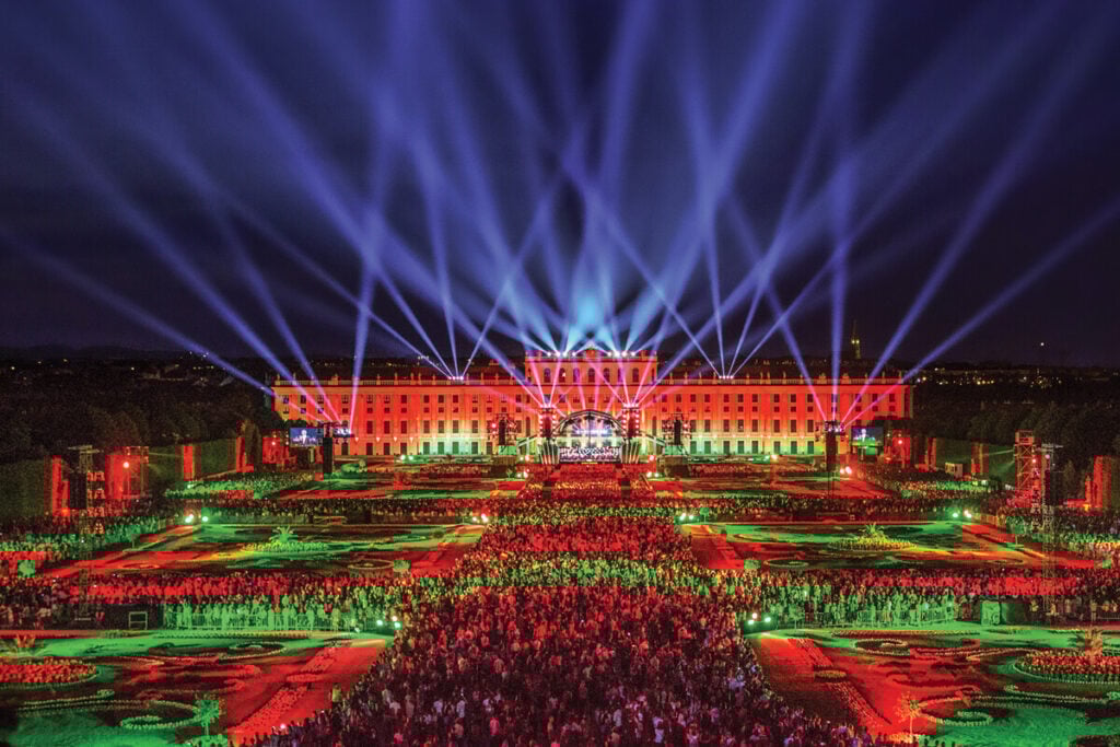 Multidão num concerto ao ar livre em frente a um palácio iluminado, com feixes de luz a cruzarem o céu nocturno. o Concerto Noturno de Verão ao ar livre, onde, todos os anos em maio ou junho, mais de 50.000 pessoas se reúnem no Palácio e Jardins de Schönbrunn, em Viena.