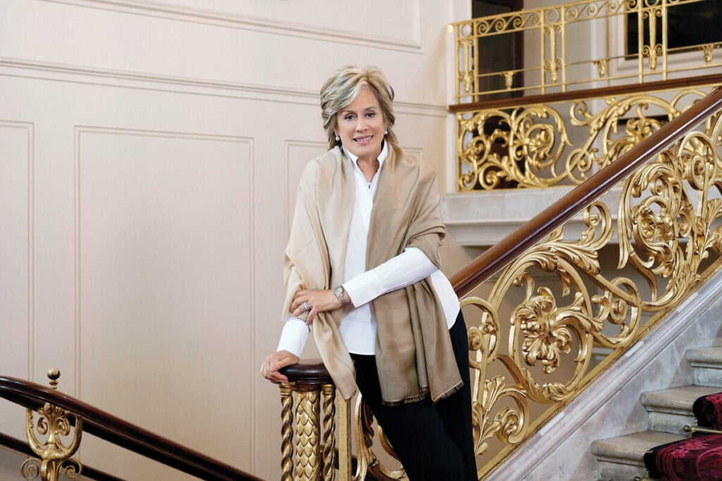 Mulher elegante encostada ao corrimão de uma escadaria com gradeamentos dourados, num interior clássico e sofisticado. Dame Kiri Te Kanawa Rolex