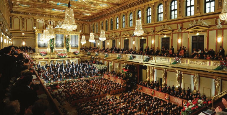 Vista geral de uma sala de concertos clássica ornamentada, com orquestra em palco, público nas galerias e grandes lustres de cristal.