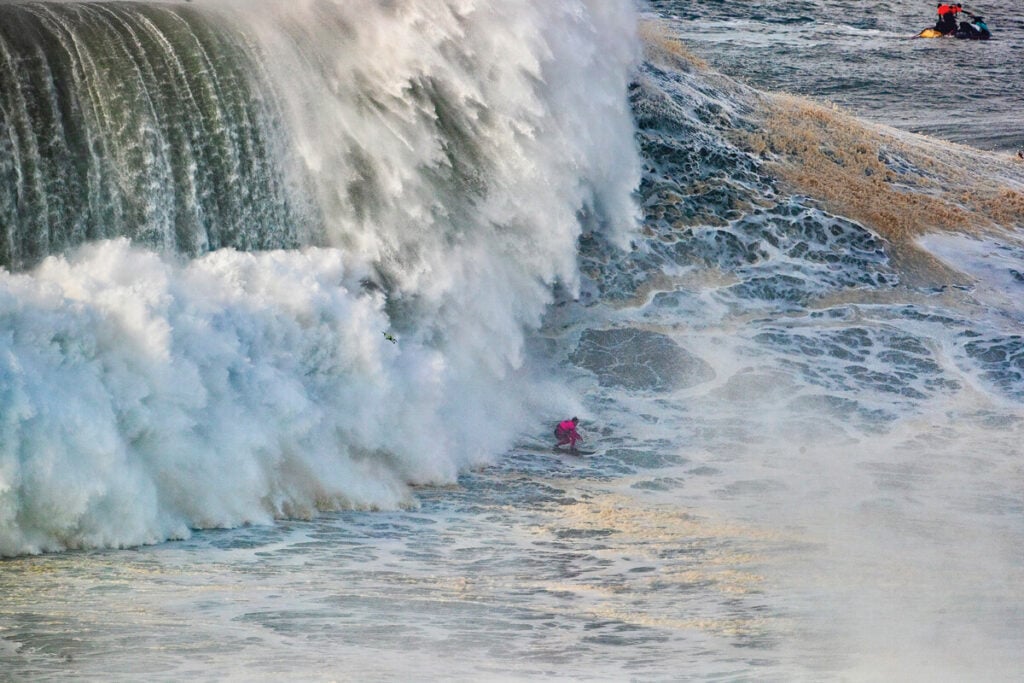 Surfista a surfar uma onda gigante em Nazaré, com a onda a erguer-se acima dele. Um barco de resgate é visível ao fundo.