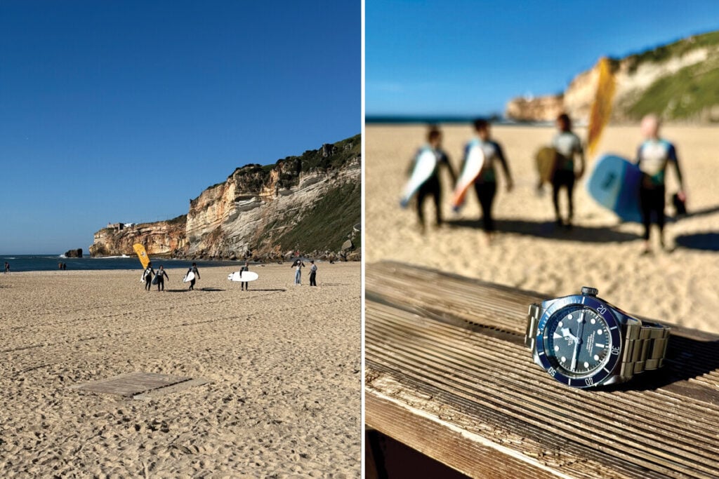 Imagem de uma praia com surfistas ao fundo e um relógio em primeiro plano. No lado esquerdo, surfistas caminham na areia com as pranchas, enquanto o relógio está em foco na madeira. O fundo está desfocado, mostrando o mar e falésias.