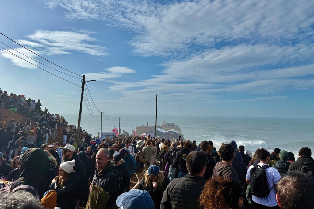 Multidão reunida nas falésias da Nazaré, assistindo a um evento de surfe. Ao fundo, é possível ver as grandes ondas e uma estrutura de apoio do evento.