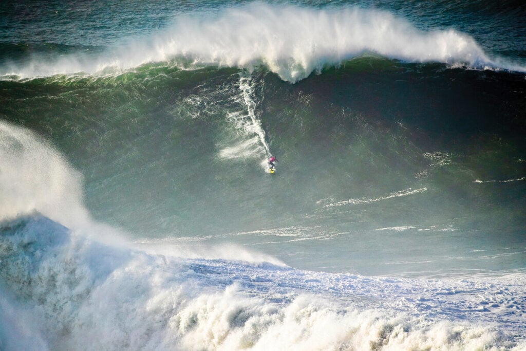 Surfista a descer a face de uma onda gigante na Nazaré, com a crista da onda a quebrar por cima.