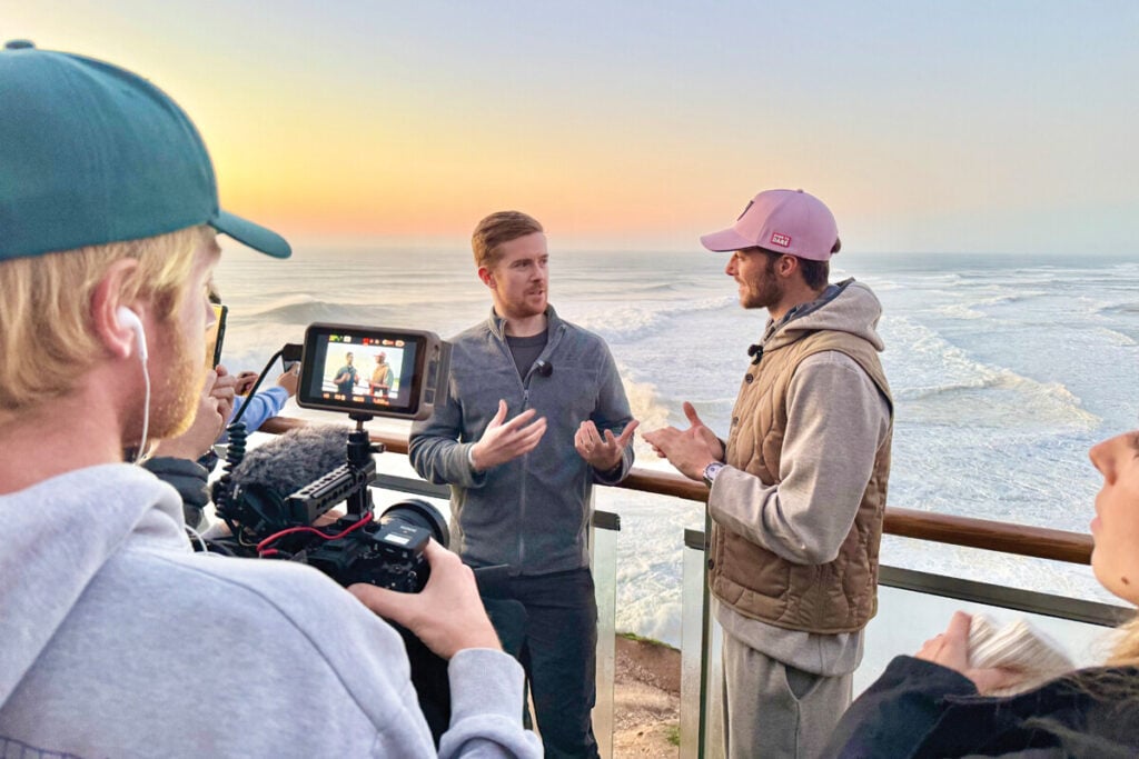 Dois homens estão a conversar enquanto são filmados por uma equipa de produção. Eles estão numa varanda com vista para o mar, com ondas grandes ao fundo. O homem à esquerda está a usar uma camisa cinza e a gesticular com as mãos, enquanto o homem à direita, com um boné rosa, está a responder. A equipa de filmagem inclui uma pessoa a manusear uma câmara, visível na imagem, e outra a gravar áudio.