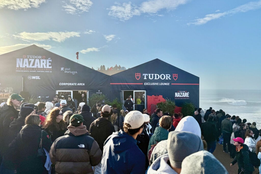 Uma multidão de pessoas reúne-se à frente de tendas com a marca "TUDOR" durante o evento "Big Wave Challenge" na Praia do Norte, em Nazaré. As tendas são identificadas como "Corporate Area" e "Exclusive Lounge". O céu está claro e há ondas visíveis ao fundo, enquanto várias pessoas estão de casacos e chapéus, algumas esperando para entrar nas tendas.