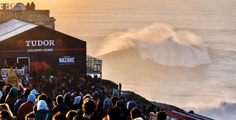 Imagem de uma multidão de pessoas em frente a uma tenda da marca Tudor, com um grande onda quebrando ao fundo. A tenda tem a inscrição "TUDOR EXCLUSIVE LOUNGE" e "TUDOR NAZARÉ". A cena ocorre ao pôr do sol, com a luz dourada iluminando a paisagem.