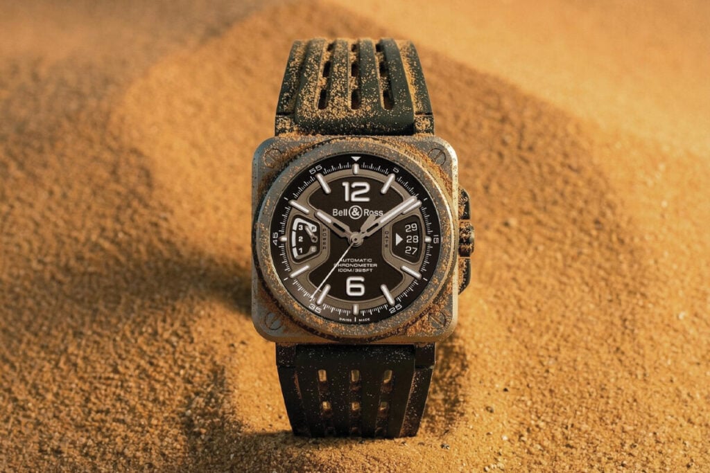 Rel&oacute;gio Bell & Ross com caixa quadrada e bracelete preta, parcialmente coberto de areia, sobre um fundo arenoso em tons dourados.
