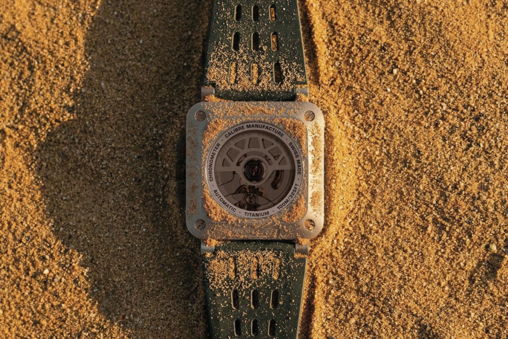 Vista traseira de um rel&oacute;gio Bell & Ross em tit&acirc;nio, com fundo transparente e bracelete verde, coberto de areia sobre um fundo arenoso.