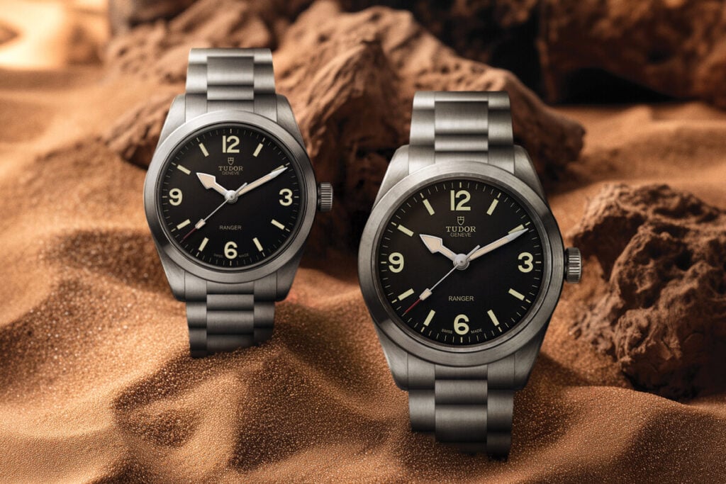 Dois rel&oacute;gios Tudor Ranger em a&ccedil;o, com mostrador preto e algarismos claros, colocados sobre areia e rochas num cen&aacute;rio des&eacute;rtico.