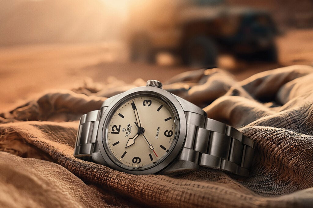 Rel&oacute;gio de pulso em a&ccedil;o com mostrador claro pousado sobre tecido, num cen&aacute;rio des&eacute;rtico ao p&ocirc;r do sol.
