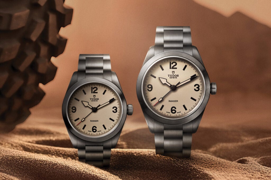 Dois rel&oacute;gios Tudor Ranger em a&ccedil;o inoxid&aacute;vel, com mostrador bege e bracelete met&aacute;lica, apresentados sobre um fundo arenoso em tons quentes.