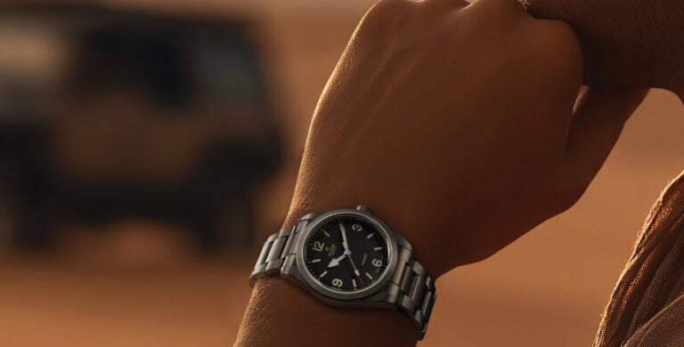 Relógio de pulso em aço no braço de uma pessoa, com mostrador preto, em ambiente desértico ao pôr do sol.