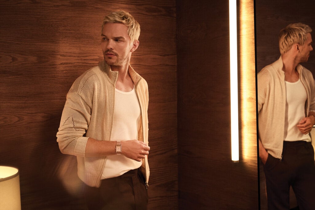 Homem loiro num espa&ccedil;o interior com paredes de madeira, a usar camisola clara e rel&oacute;gio no pulso, junto a um espelho iluminado.