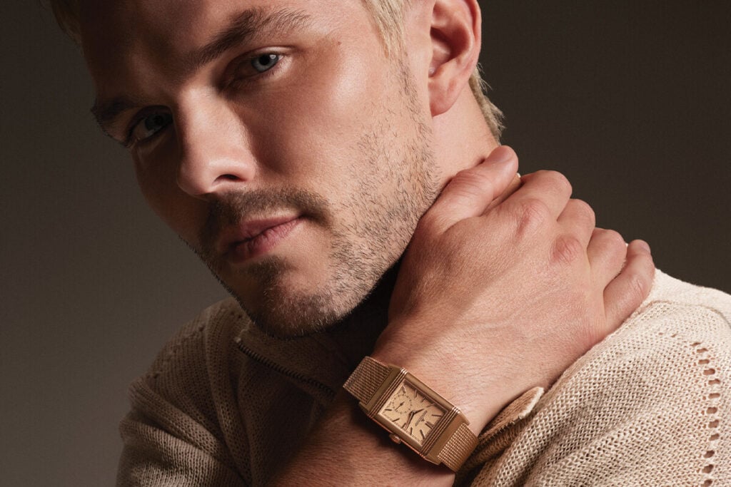 Close-up de um homem com barba curta a usar um rel&oacute;gio dourado de caixa rectangular no pulso, com a m&atilde;o junto ao pesco&ccedil;o.