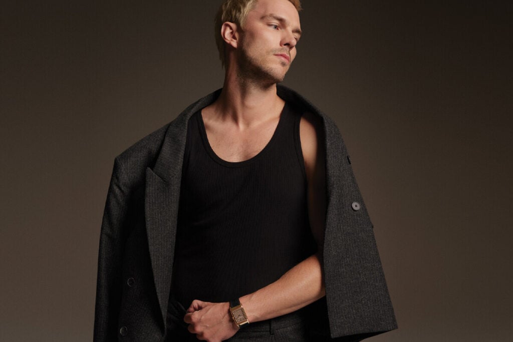 Homem em est&uacute;dio, de perfil, a usar camisola preta sem mangas, casaco cinzento pousado sobre os ombros e rel&oacute;gio no pulso, sobre fundo neutro.