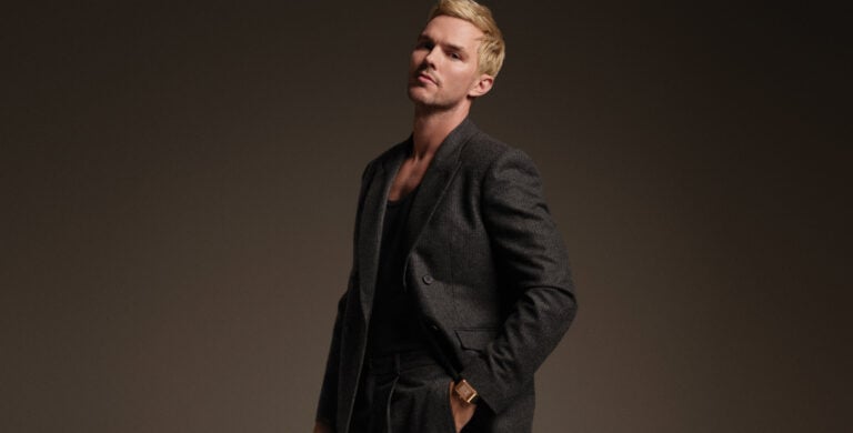 Homem loiro, de fato escuro, posa em estúdio com a mão no bolso, usando um relógio dourado no pulso.