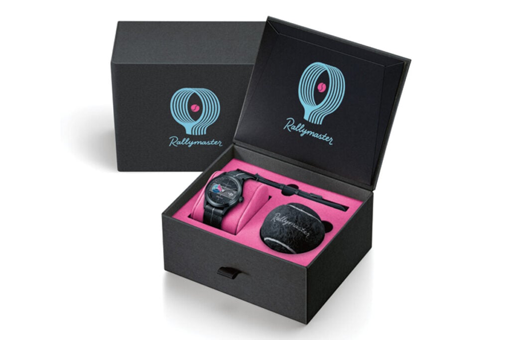 Imagem de uma caixa de apresentação do relógio Maurice de Mauriac modelo Rallymaster. A caixa contém o relógio, uma bola de ténis e uma ferramenta, tudo envolto em material acolchoado rosa. O logotipo "Rallymaster" está visível na parte superior da caixa e na bola de ténis.