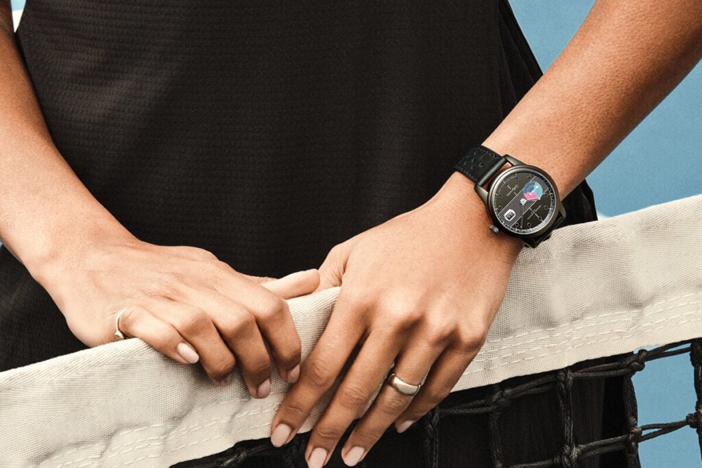 Imagem de uma mão segurando a rede de um campo, com um relógio Maurice de Mauriac modelo Rallymaster visível no pulso. O relógio tem uma caixa preta e mostrador com gráficos coloridos, e a correia é preta, de couro perfurado.