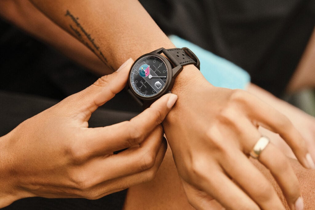 Imagem de uma mão ajustando um relógio Maurice de Mauriac modelo Rallymaster no pulso, com o mostrador preto e detalhes coloridos visíveis. O fundo é suave, com foco no relógio e na mão.