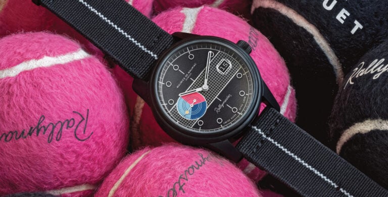 Imagem de um relógio Maurice de Mauriac modelo Rallymaster, posicionado sobre várias bolas de ténis de cores rosa e preta. O relógio tem uma caixa preta e mostrador com detalhes gráficos, incluindo círculos coloridos em tons de azul e vermelho, e a correia é de nylon preto com riscas brancas.