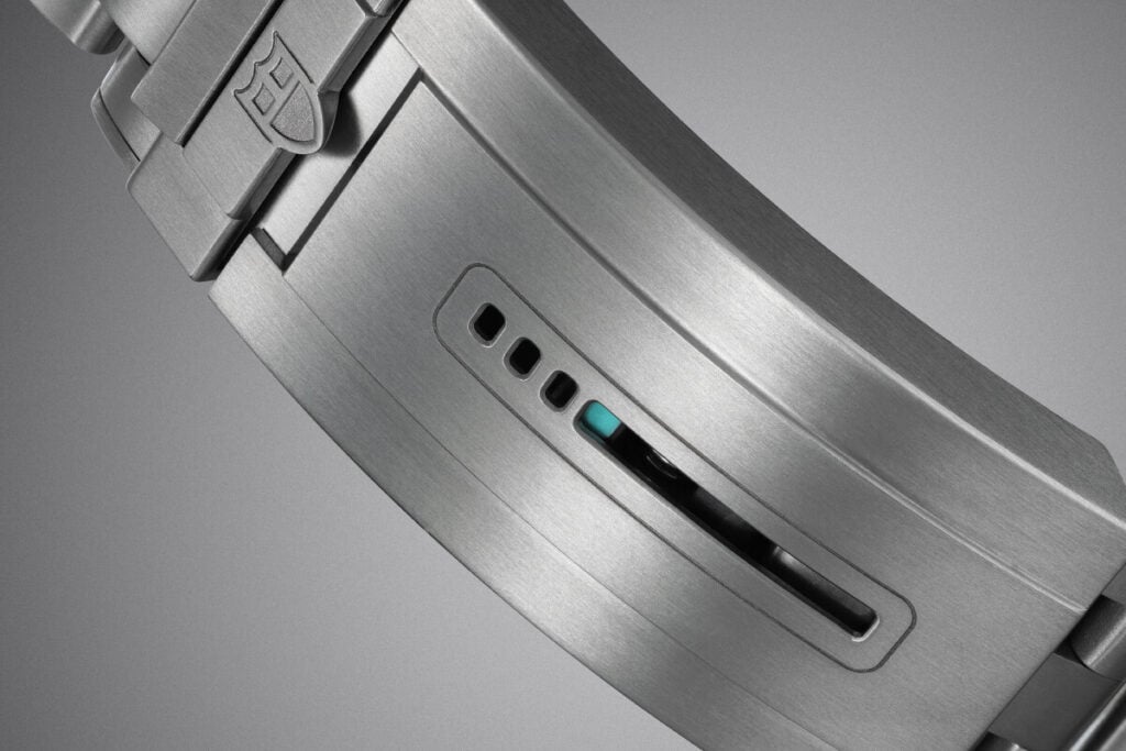 Fecho met&aacute;lico de rel&oacute;gio com acabamento escovado, mostrando detalhes do mecanismo de ajuste. O fecho &eacute; gravado com o logo da marca Tudor e possui um indicativo azul, com um sistema de fivela e orif&iacute;cios para ajuste.