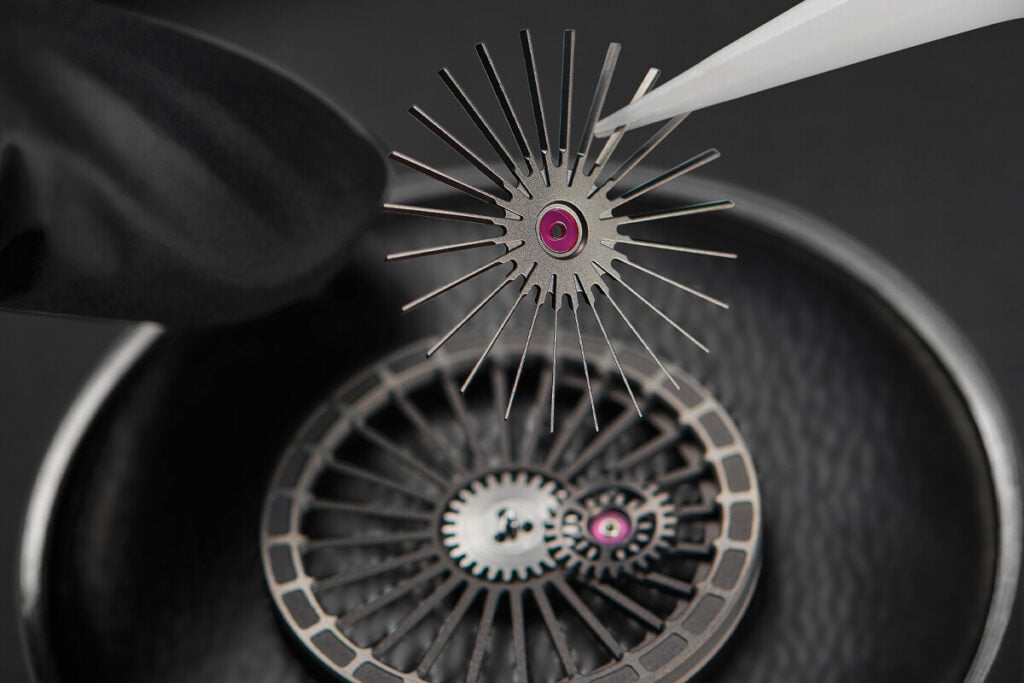 Close-up de componentes do movimento de um rel&oacute;gio, com uma pe&ccedil;a central em forma de estrela sendo cuidadosamente manuseada e lubrificada.