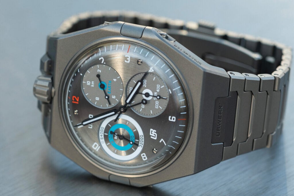 Rel&oacute;gio de pulso Urwerk em tit&acirc;nio com mostrador cinzento, tr&ecirc;s submostradores e detalhes em azul, apresentado sobre uma superf&iacute;cie lisa.