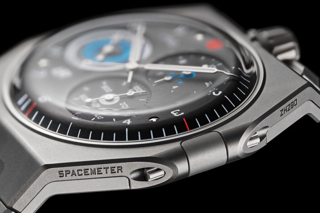 Close-up de um rel&oacute;gio moderno, com caixa em tit&acirc;nio, mostrador detalhado e marcadores de tempo precisos. O modelo exibe complica&ccedil;&otilde;es de medi&ccedil;&atilde;o e possui detalhes em azul, vermelho e prata.