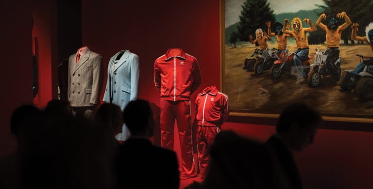 Exposição num museu com fatos e fatos de treino sem manequins, alinhados numa sala vermelha, ao lado de um quadro com figuras mascaradas em motas, observados por visitantes em silhueta.