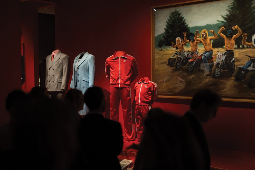 Exposi&ccedil;&atilde;o num museu com fatos e fatos de treino sem manequins, alinhados numa sala vermelha, ao lado de um quadro com figuras mascaradas em motas, observados por visitantes em silhueta.