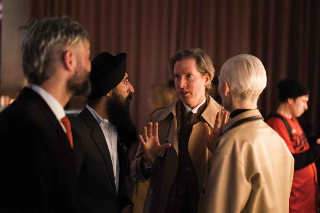 Grupo de visitantes conversa numa sala de museu; um homem ao centro gesticula enquanto fala com outras tr&ecirc;s pessoas, num ambiente interior com luz quente.