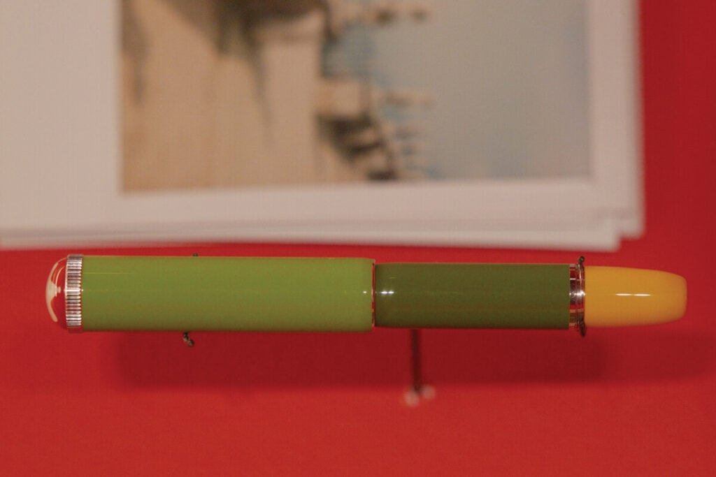 Caneta estilogr&aacute;fica de cor verde, com extremidade amarela, apresentada na horizontal sobre um fundo vermelho.