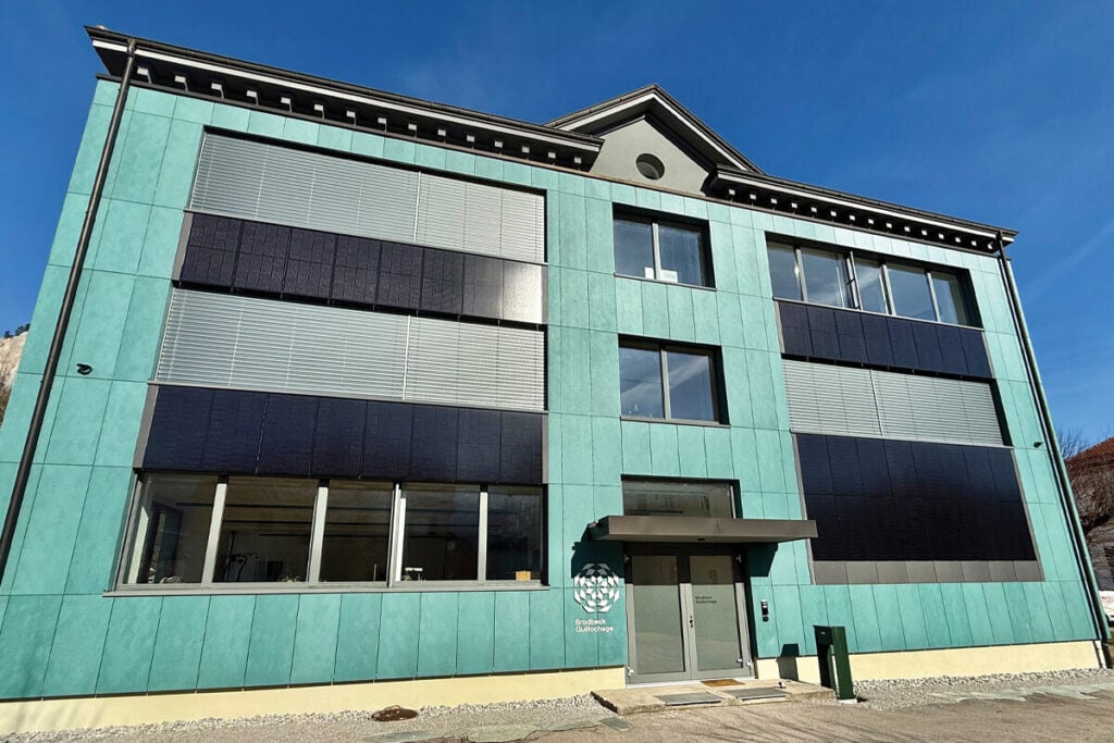 Fachada de um edifício moderno com revestimento verde-água e painéis solares integrados nas janelas, sob céu azul.