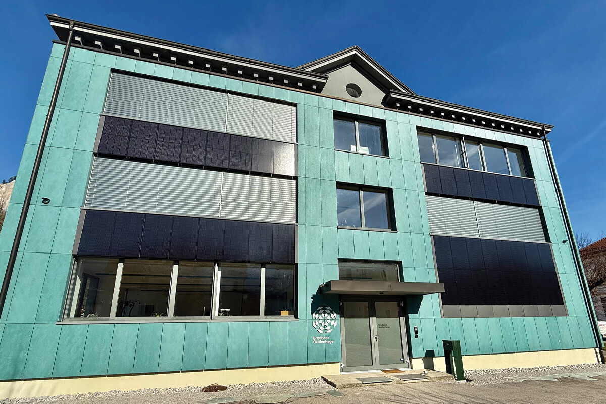 Fachada de um edifício moderno com revestimento verde-água e painéis solares integrados nas janelas, sob céu azul.