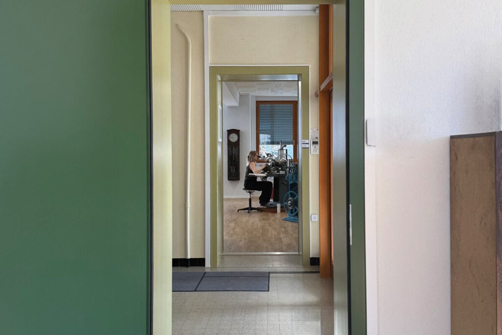 Vista de um corredor interior que conduz a uma sala onde uma pessoa trabalha numa máquina de precisão.