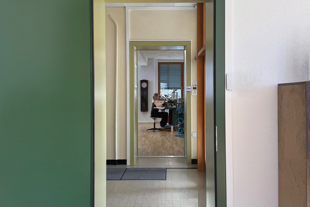 Vista de um corredor interior que conduz a uma sala onde uma pessoa trabalha numa máquina de precisão.