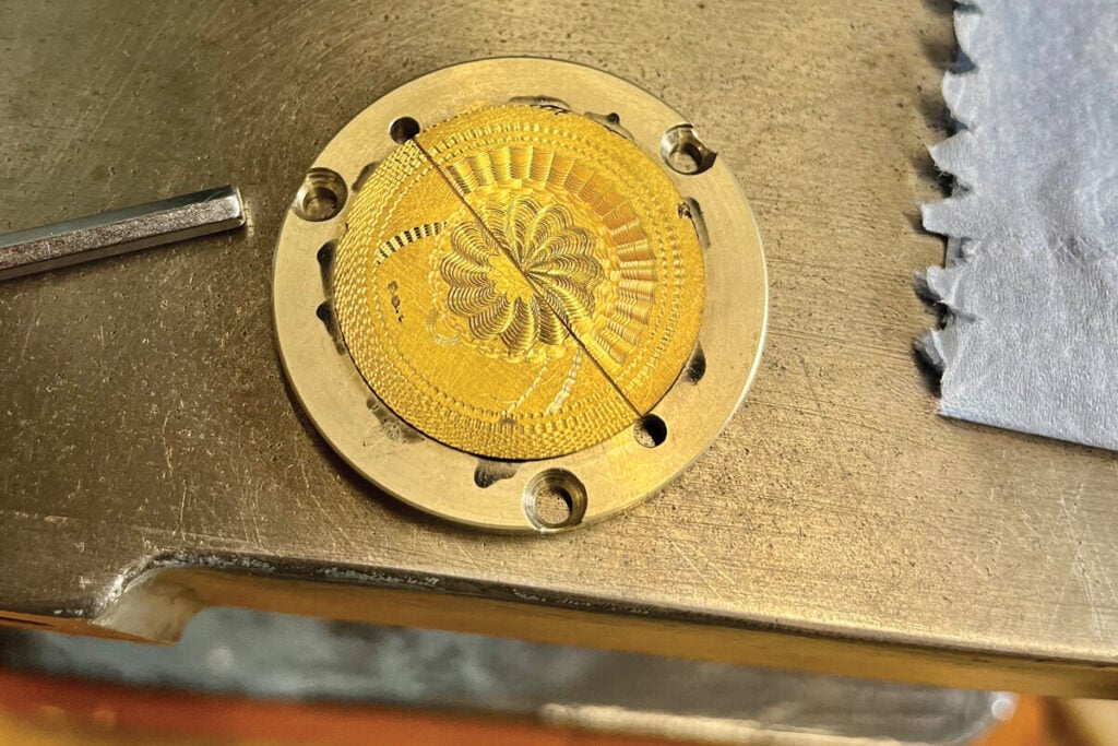 Disco metálico dourado com padrão decorativo gravado, fixo numa base de máquina, com uma linha diagonal marcada sobre a superfície.