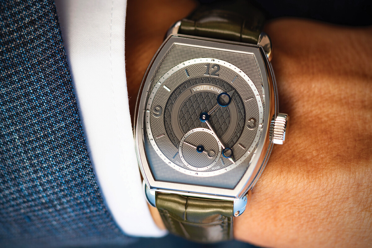 Relógio de pulso de luxo com caixa prateada e bracelete em pele verde, usado no pulso de uma pessoa com fato azul.