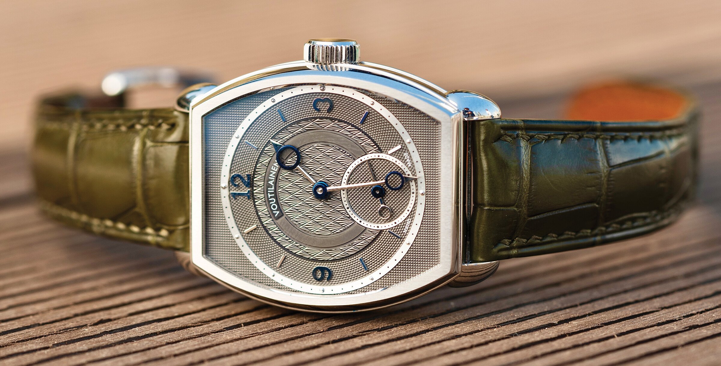 Rel&oacute;gio de pulso de luxo com caixa met&aacute;lica prateada e bracelete em pele verde, colocado sobre uma superf&iacute;cie de madeira.