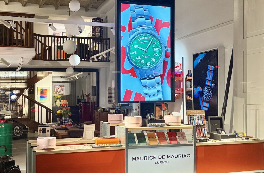 Montra da boutique Maurice de Mauriac em Zurique com ecrã digital de relógio. Vista noturna da fachada da boutique Maurice de Mauriac, localizada na Rämistrasse 5, em Zurique. A montra iluminada exibe relógios e um grande ecrã digital com um modelo da marca. Ao lado direito encontra-se o espaço da casa de leilões Koller Auktionen. Um homem entra no edifício pela porta lateral, enquanto o interior moderno da loja é visível através das grandes janelas. Foto: Miguel Seabra/Espiral do Tempo