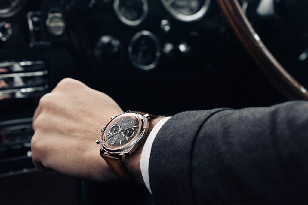relógio no pulso dentro de carro três relógio em assento preto mão ao volante com Breitling Top Time B01 Chronograph 41 Tribute to Aston Martin DB5 1 | Foto: Breitling